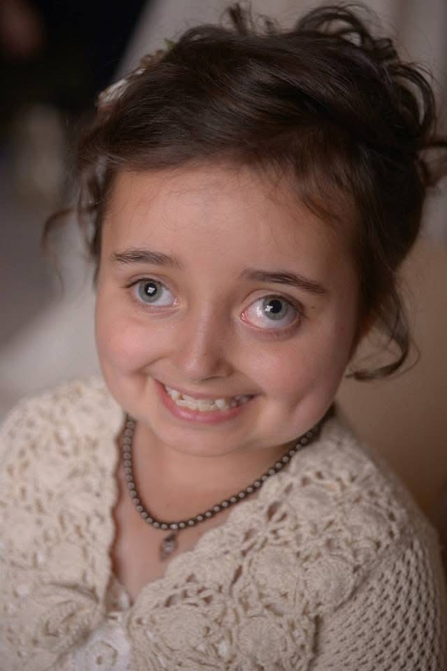 Julia Gebert smiles warmly at the camera in a close-up portrait taken when she was a young girl. She has large green eyes, rosy cheeks, and deep dimples that brighten her joyful smile. Her dark brown hair is softly gathered into a loose updo, with wisps of hair framing her face and lending the portrait a natural, playful elegance.